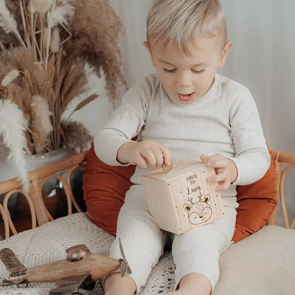 Kind sitzt im Kinderzimmer und wirft Münze in personalisierte Holz-Spardose mit Reh-Motiv und eigenem Namen